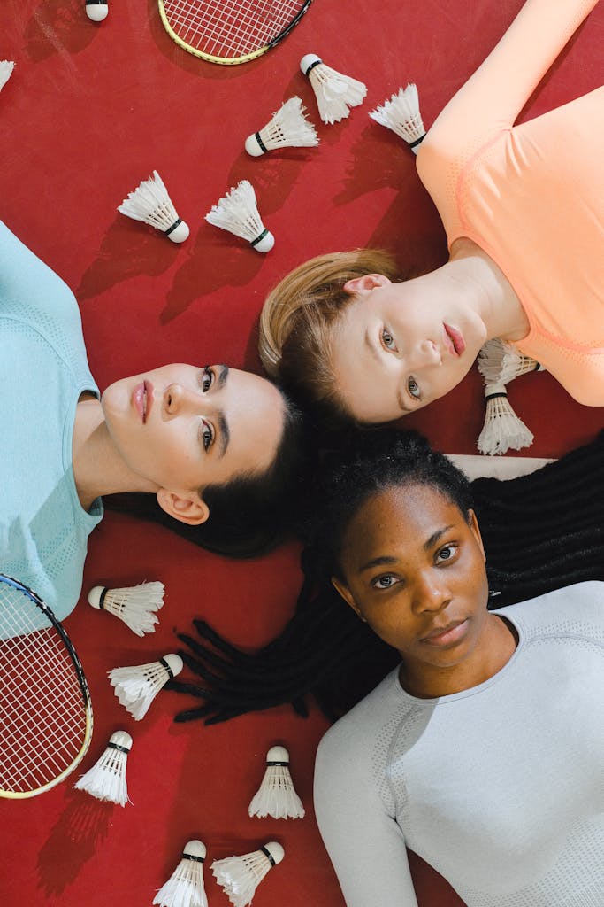 Three women in sportswear lying on a badminton court, surrounded by shuttlecocks.