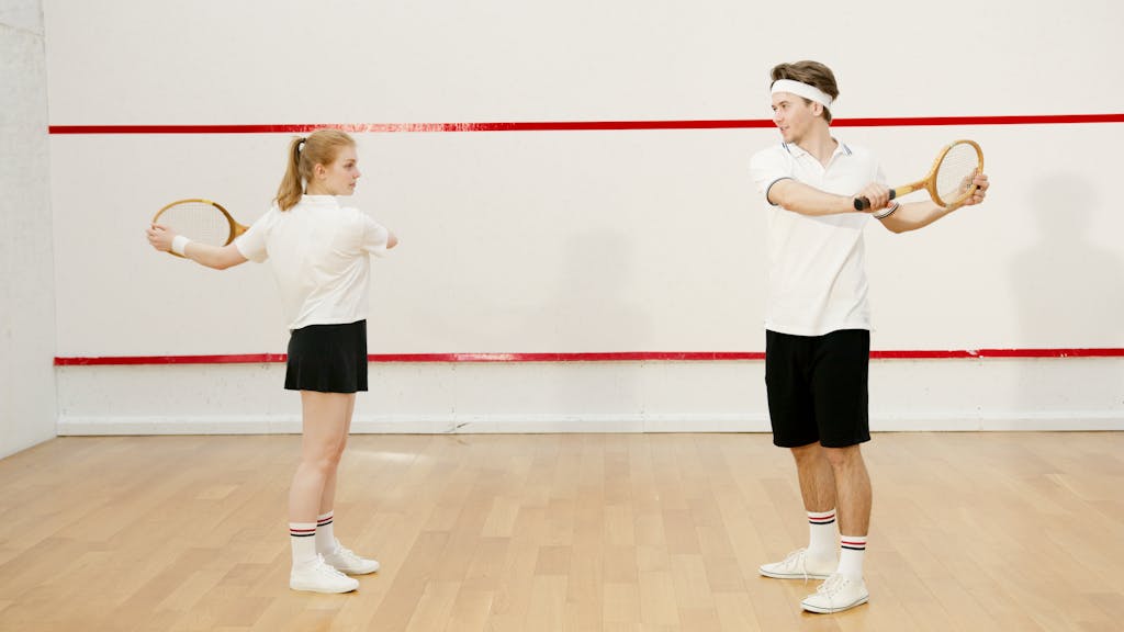 Two young squash players warming up with rackets in a squash court.