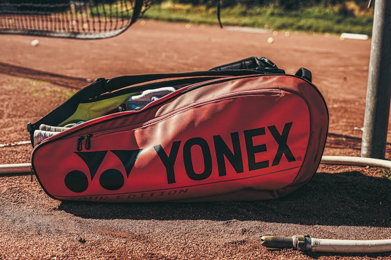 Close-up of a Yonex tennis bag lying on a clay court under sunlight, showcasing sports gear.