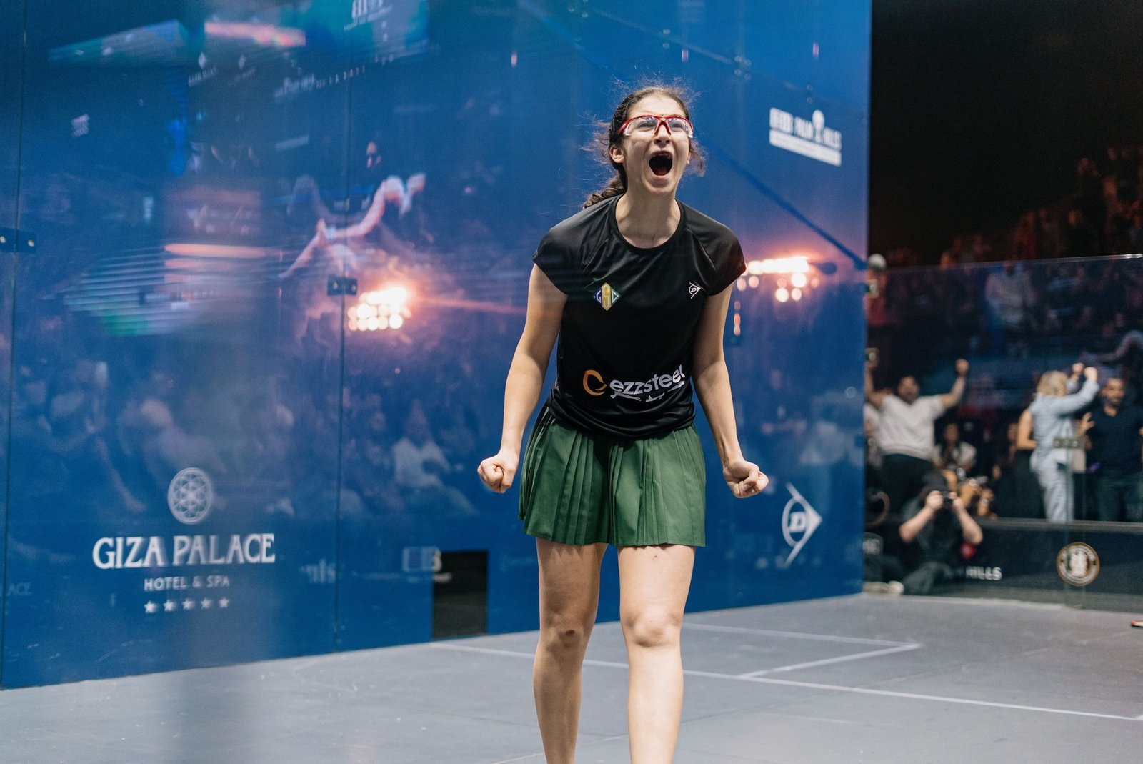 Amina Orfi screaming in celebration in front of the blue glass wall of the show court in Giza after becoming the youngest women's World Champion in history