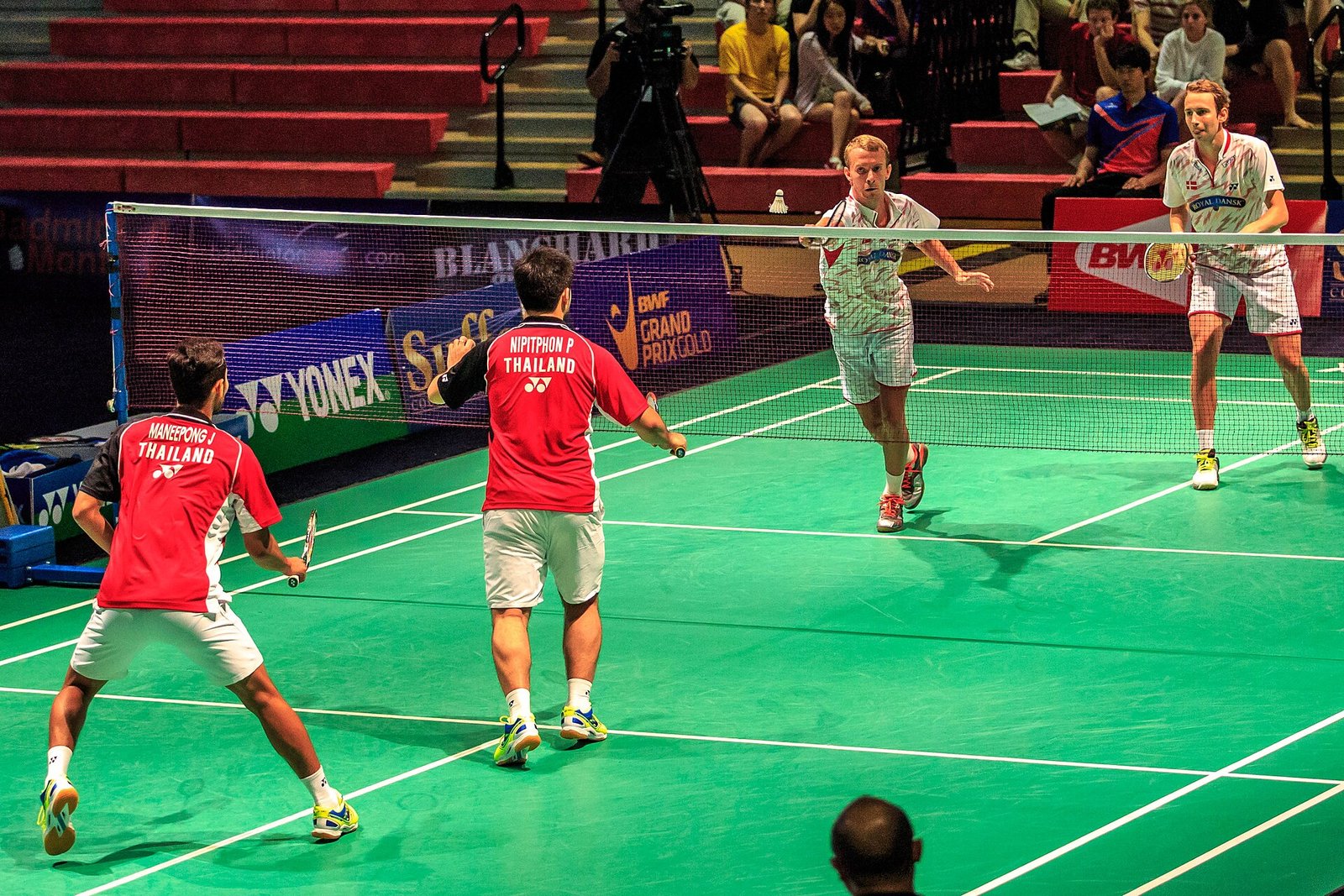 A doubles rally in progress with four players on court, one pair formed up front-and-back in attack and the shuttle crossing low over the net
