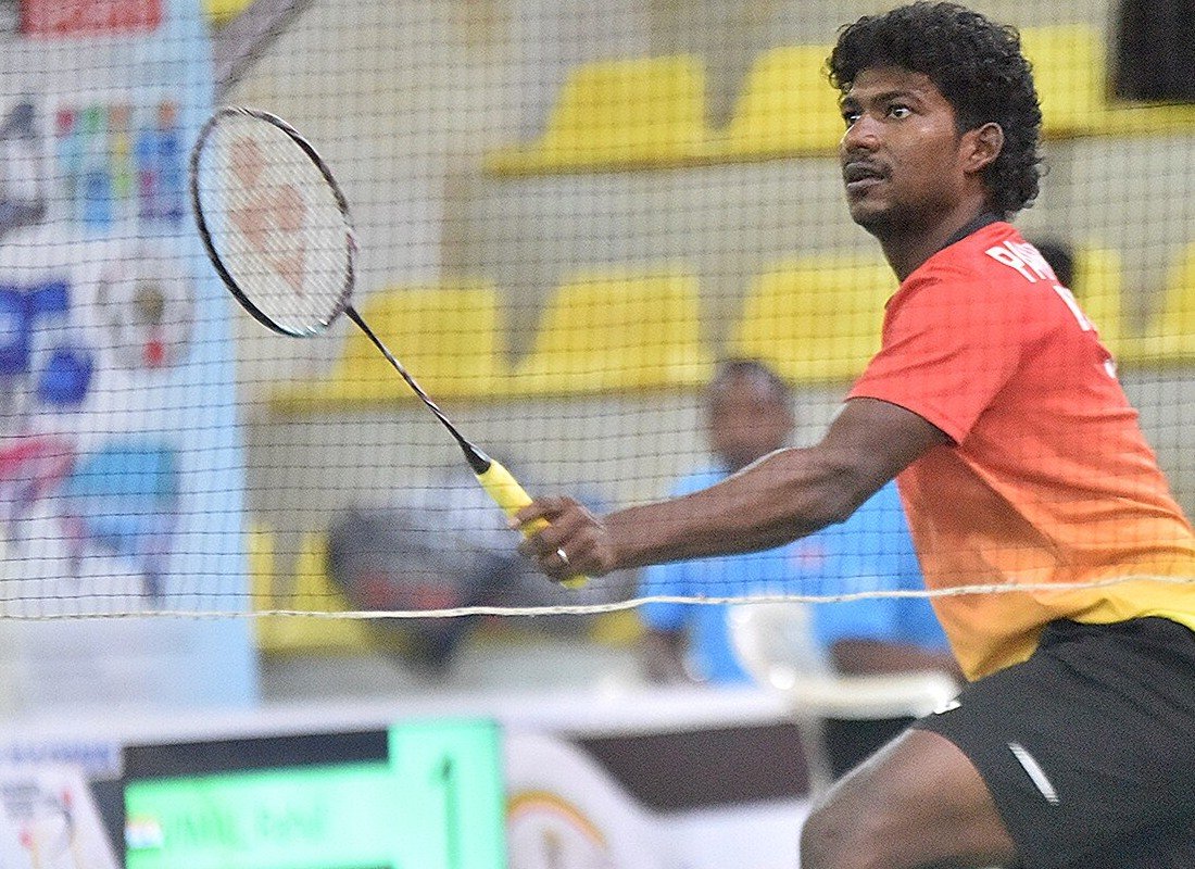 A player's hand gripping a badminton racket handle during a rally, fingers wrapped around the grip with the forearm extended and the racket head raised in front of the body