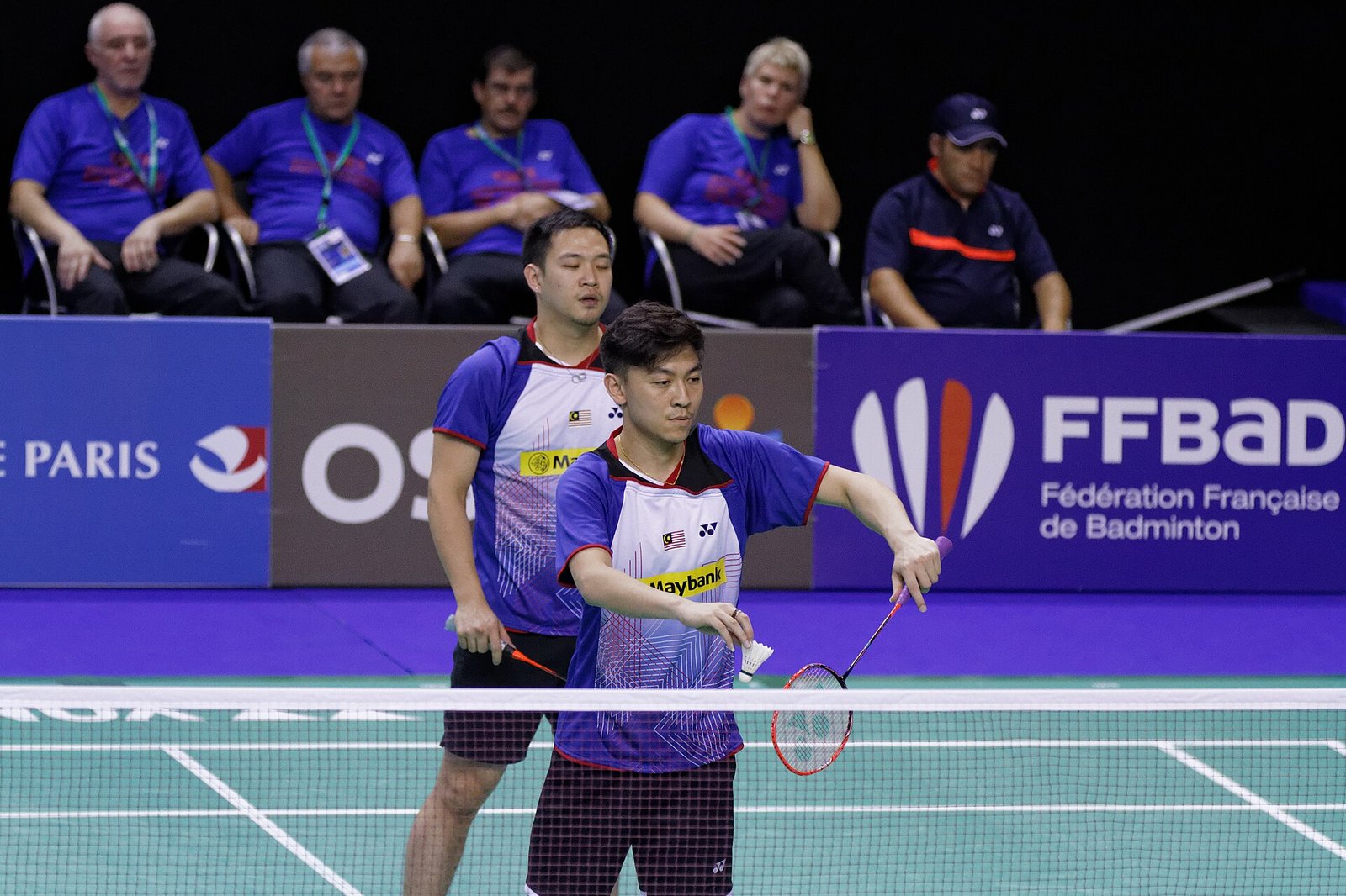 A doubles player about to serve at the net, holding the shuttle in front of his racket with the racket held low, while his partner waits behind him in the ready position