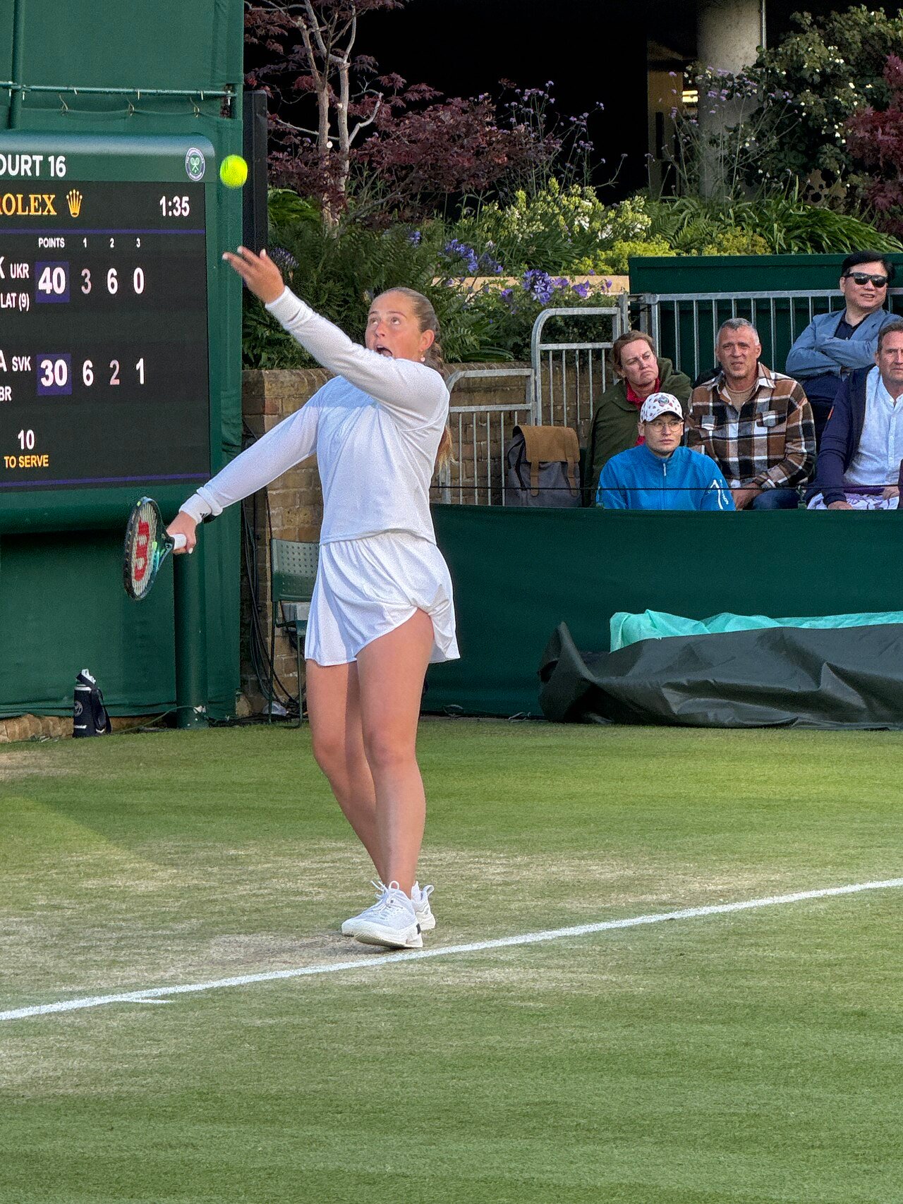 A woman tennis player at the top of her service motion, racket raised behind the tossed ball