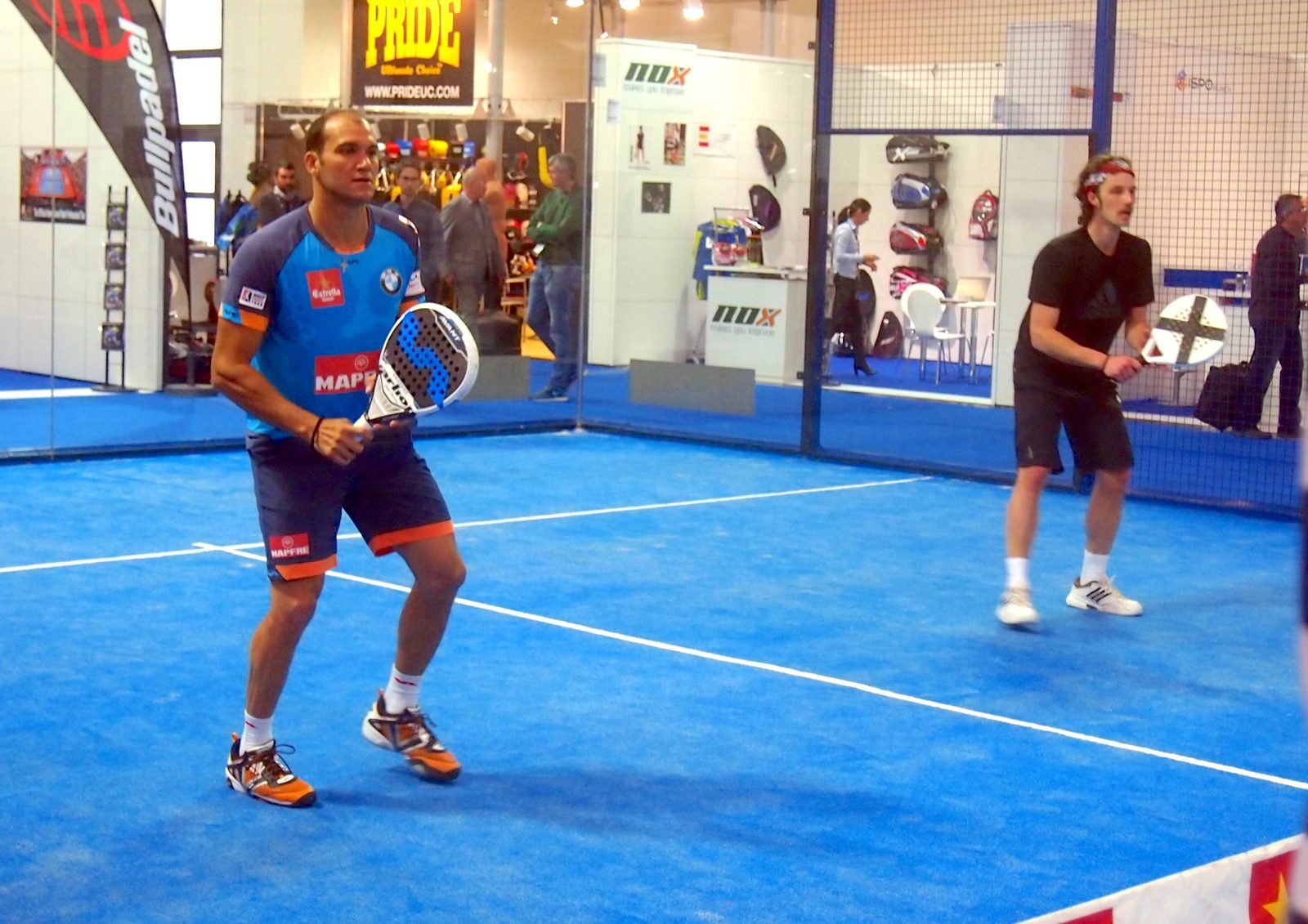 Indoor padel court inside a large arena hall with seating around the glass walls