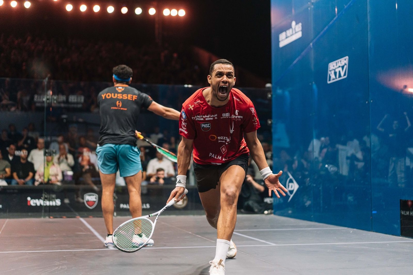 Mostafa Asal roaring in celebration on the glass court in Giza after winning the 2026 PSA World Championship final, with Youssef Ibrahim walking away behind him