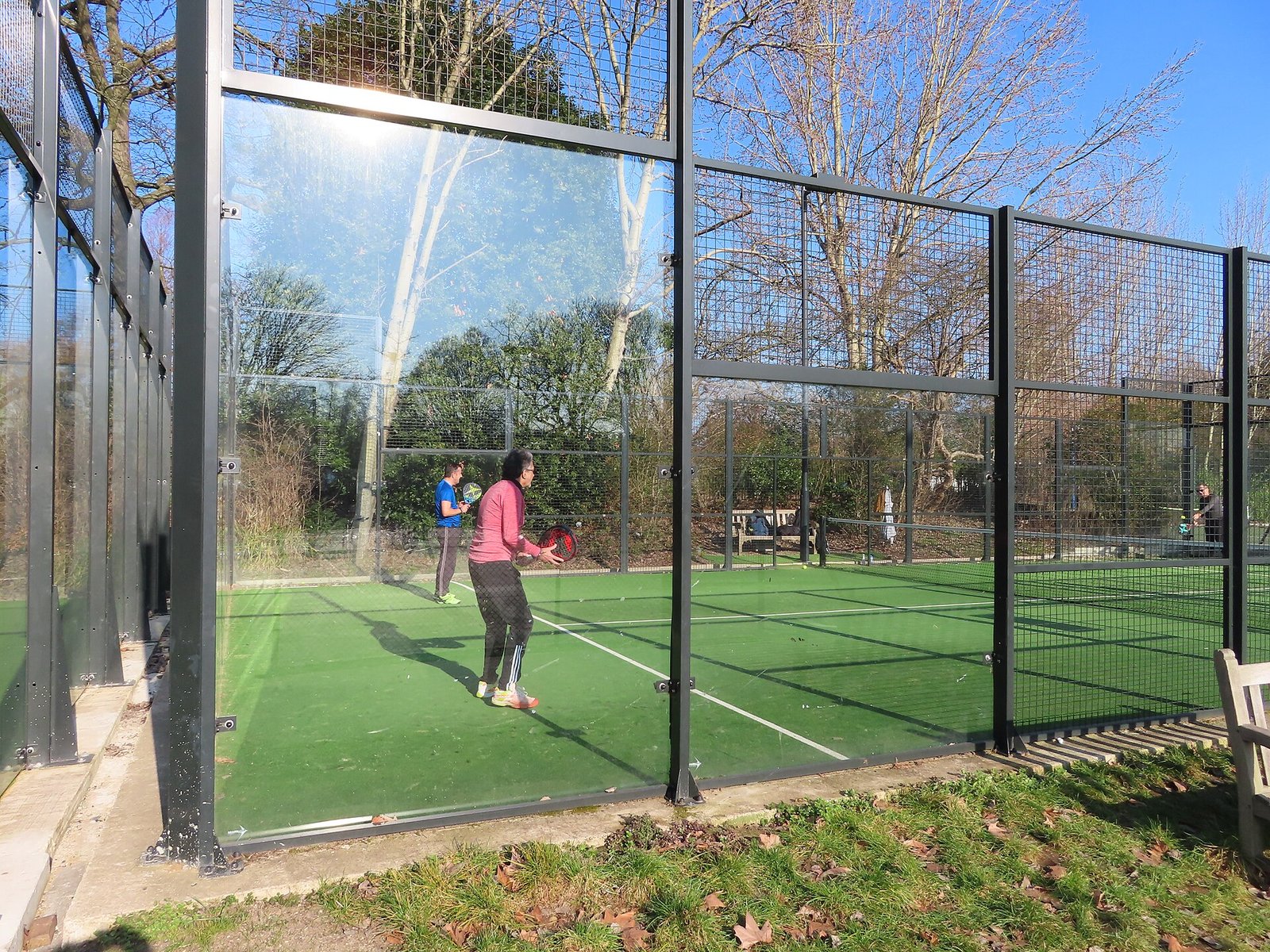 A padel pair playing inside a glass-walled court seen through the back glass, with the mesh fencing above the glass and the service lines marked on the green court