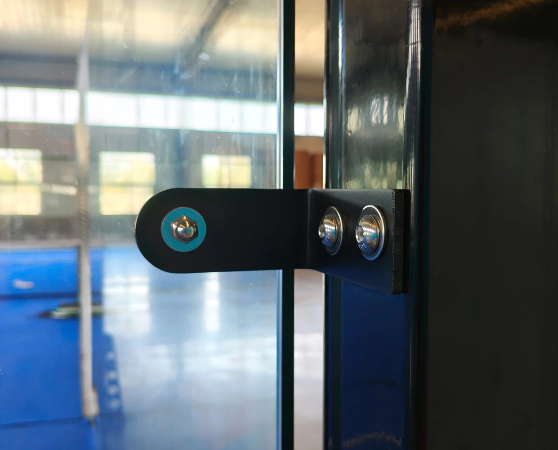 Close-up of the bolted steel fitting joining two 12mm tempered glass panels on a panoramic padel court