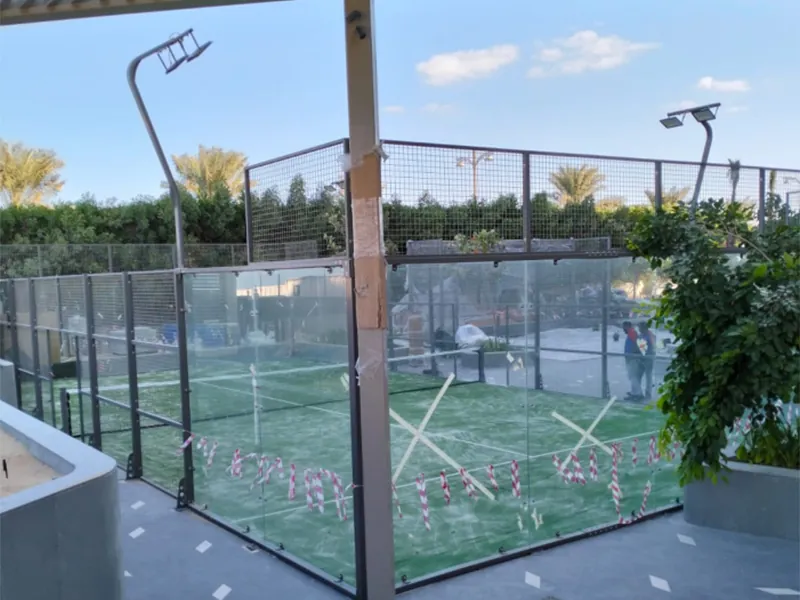 Tempered glass panels being installed on an outdoor padel court, with floodlight poles already in place