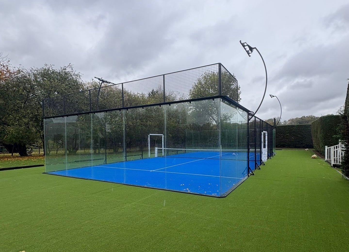 A panoramic padel court with full-height blue-framed glass back walls and green playing surface, seen empty from behind one baseline