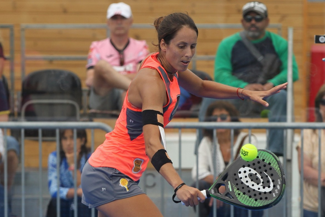 A padel player stretched low into a forehand during a rally on an indoor court, ball just leaving the perforated racket face