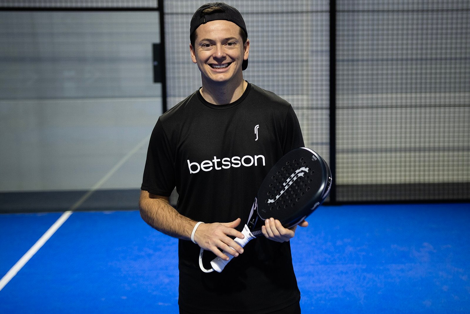 Padel player holding a perforated padel racket with wrist strap in front of the glass wall of an indoor court