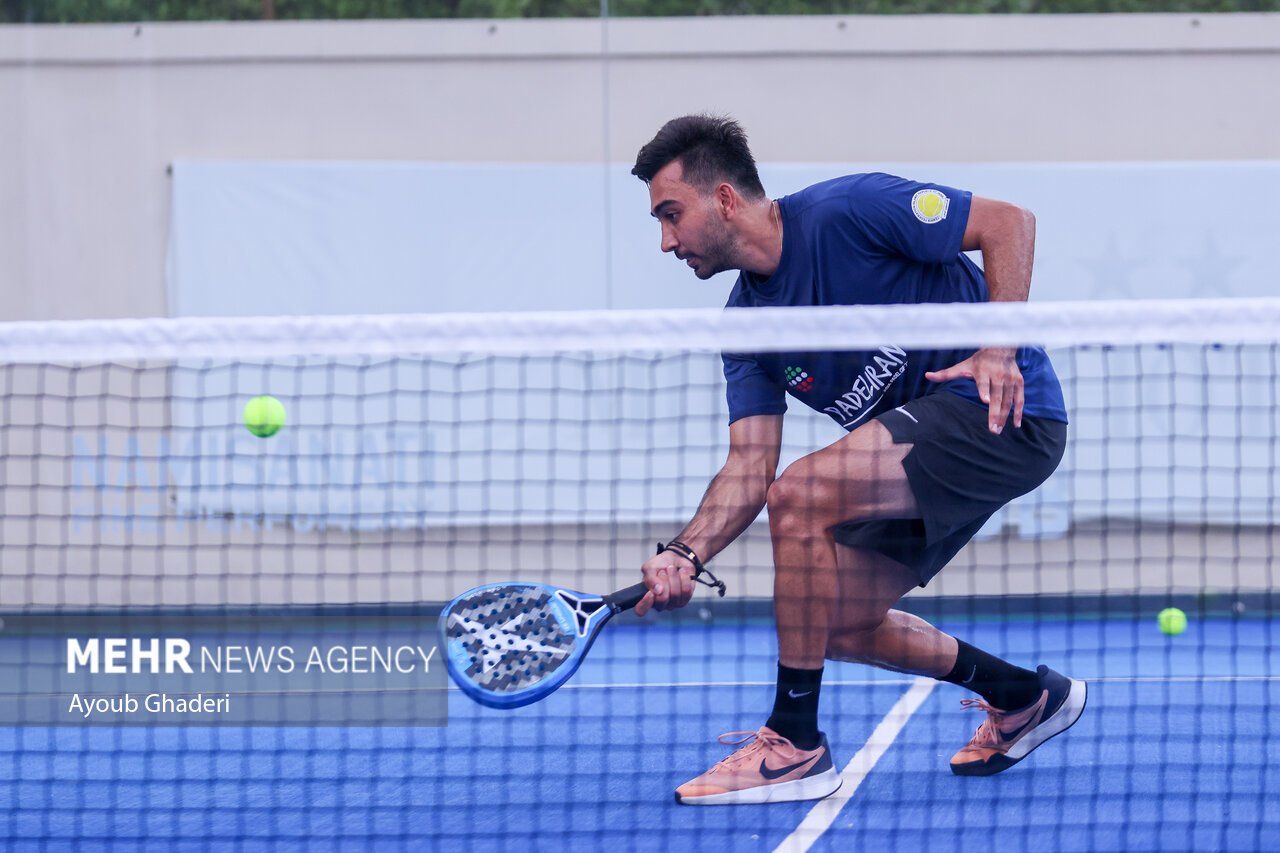A padel player crouched low at the net playing a backhand volley, photographed through the net from the opposite side of the court