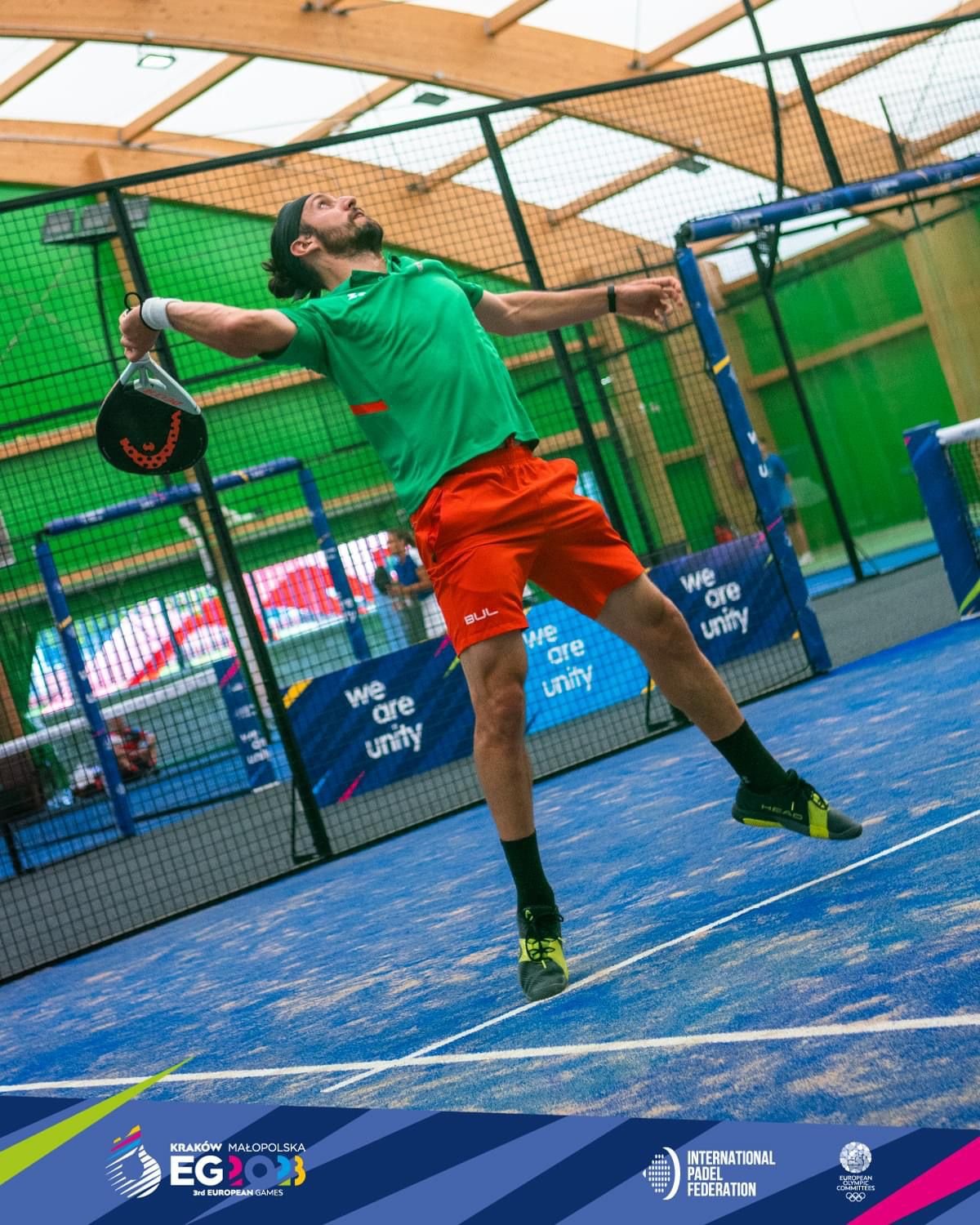 A padel player stretched into the high overhead position for a bandeja, racket cocked behind his head and eyes on the ball during a match