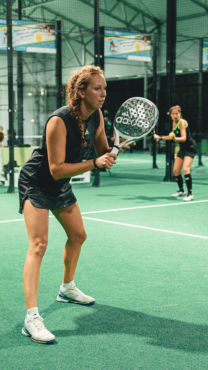 A padel player in a low ready position with her racket up in front of her, set to receive during a rally on an indoor court while her partner waits behind