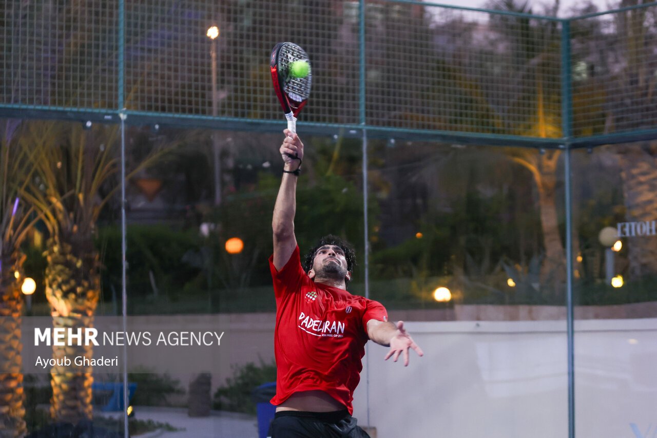 A padel player at full stretch overhead making contact with the ball for a smash on an outdoor glass court at dusk