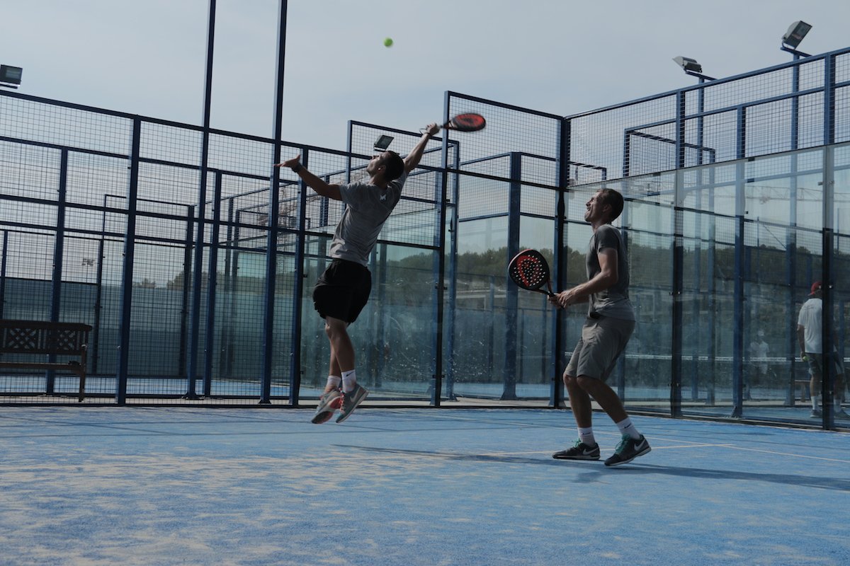 A padel player leaping at the back of the court to smash the ball down off a lob while his partner watches, the glass and mesh enclosure behind them