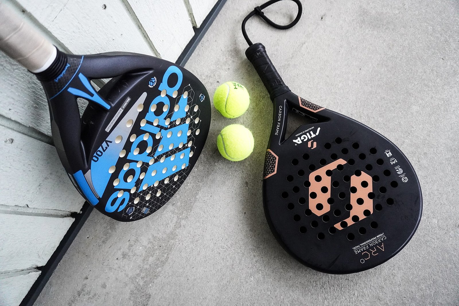 Two padel rackets and two padel balls resting on court flooring, one racket with its wrist safety cord looped beside it