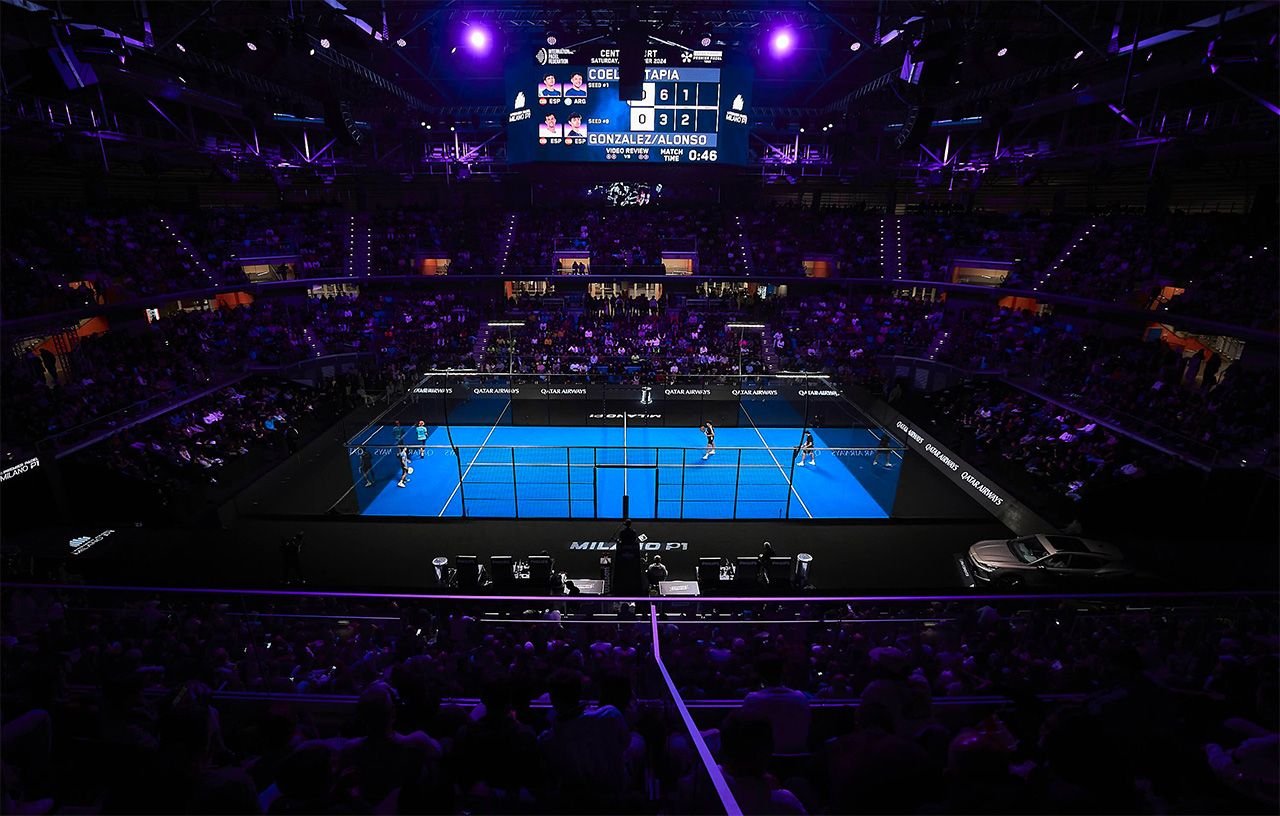 A Premier Padel centre court inside a packed indoor arena during a night session, the scoreboard glowing above the blue glass court