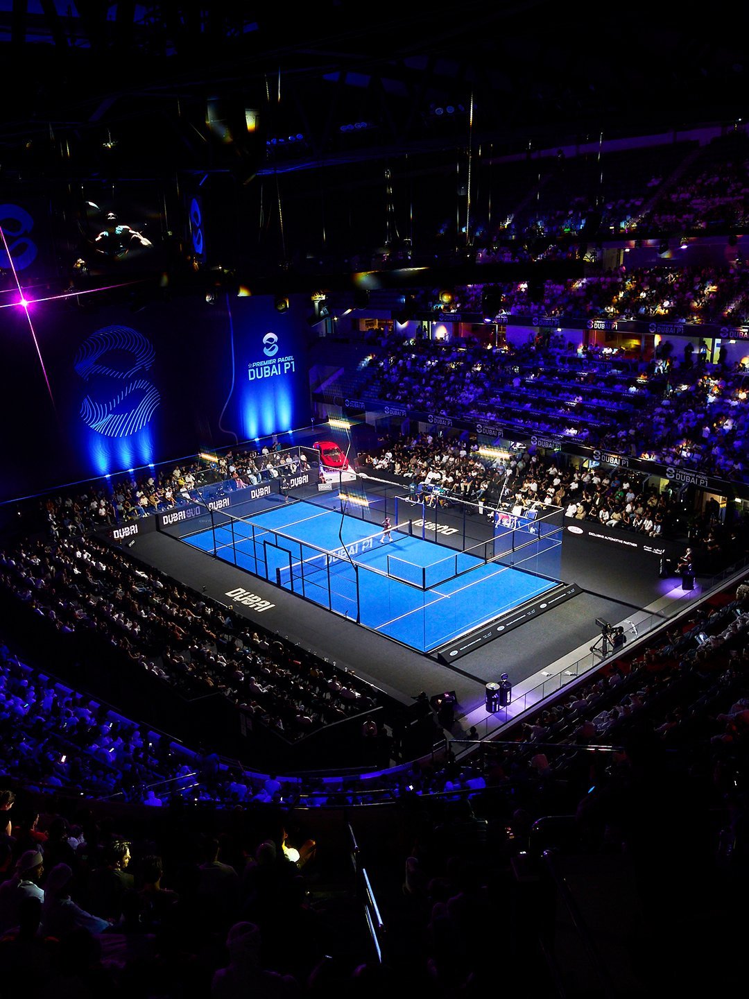 The blue glass show court at the Dubai Premier Padel P1 glowing under arena lights, surrounded by a full crowd in the darkened stands