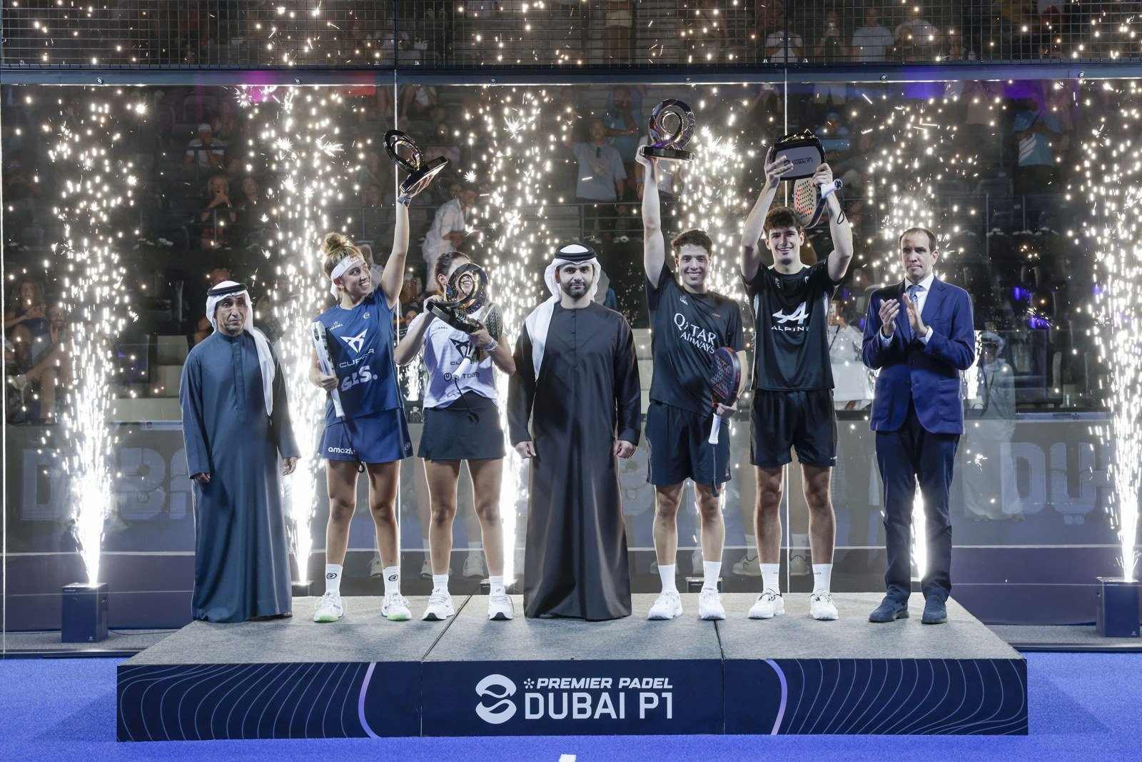 The men's and women's champions on the podium at the Dubai Premier Padel P1, trophies raised as fireworks fire behind the glass wall