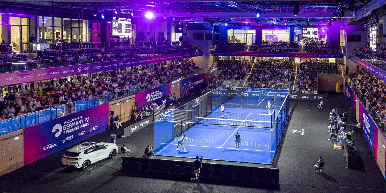 A women's doubles match in play on the glass show court at the Germany P2 in Düsseldorf, with tiered seating rising on both sides of the court