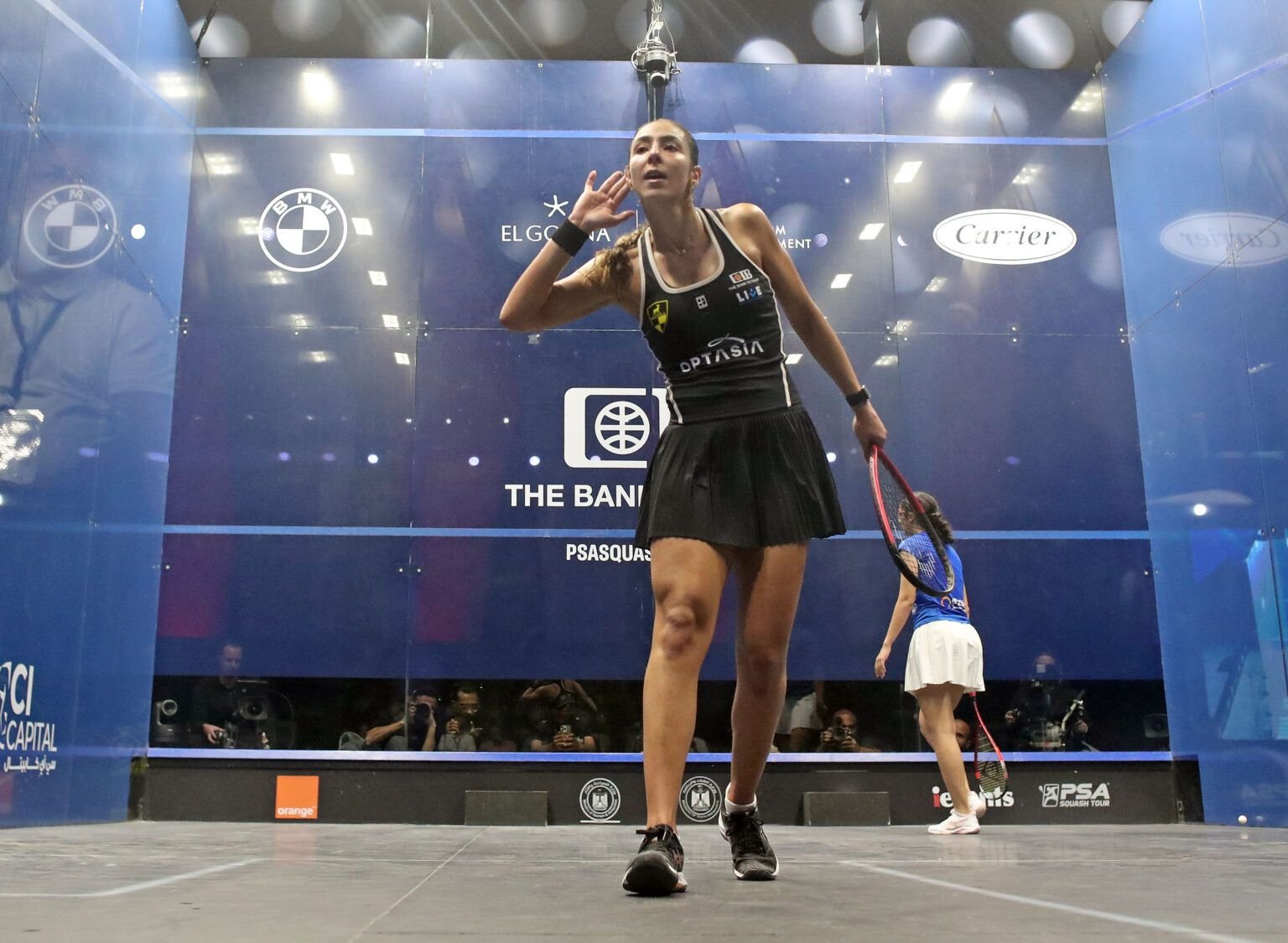 A professional squash player on an all-glass PSA Tour show court during a rally, with the crowd and cameras visible behind the glass back wall