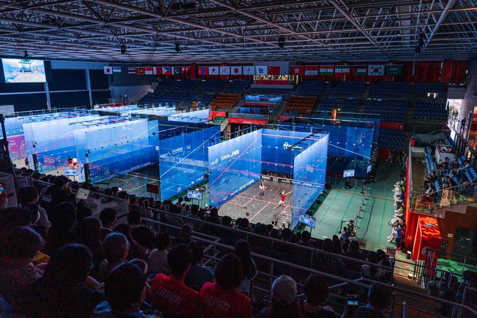 A hall of all-glass squash courts under arena lights at the Aichi-Nagoya 2026 Asian Games, with spectators watching from the stands