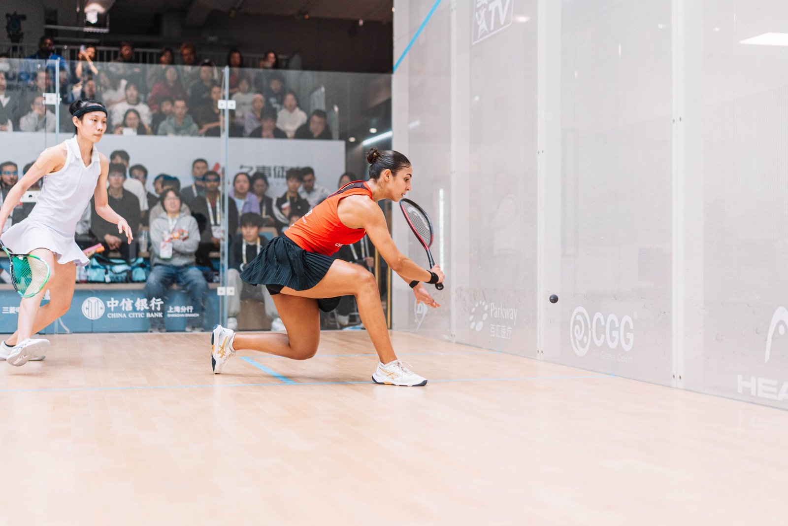 A squash player stretched low in the front corner digging out a ball that has come off the side wall close to the front wall
