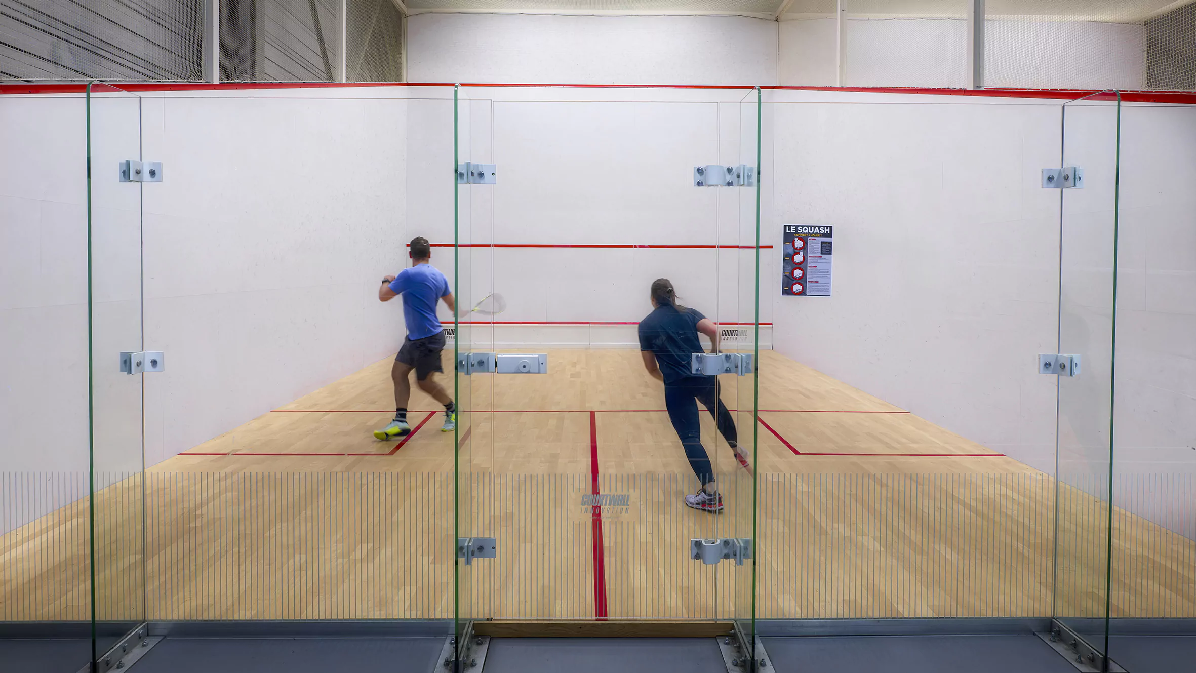 Players contesting a rally on an indoor squash court seen through the glass back wall — the front wall, tin and floor markings visible ahead of them
