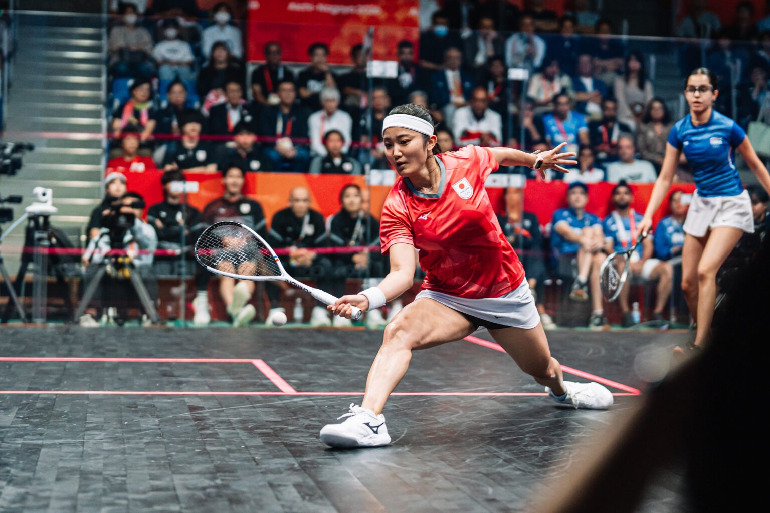 A squash player in a deep lunge at the front of the court playing a low forehand with the ball just off the floor close to her racket
