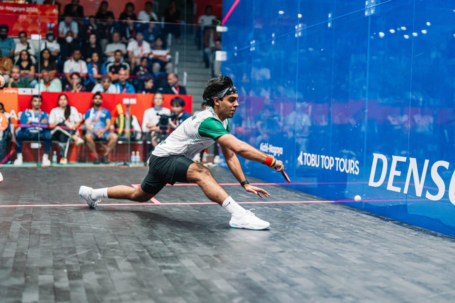 A men's squash player stretched at full length along the blue glass side wall, reaching a ball during a match at the Aichi-Nagoya 2026 Asian Games