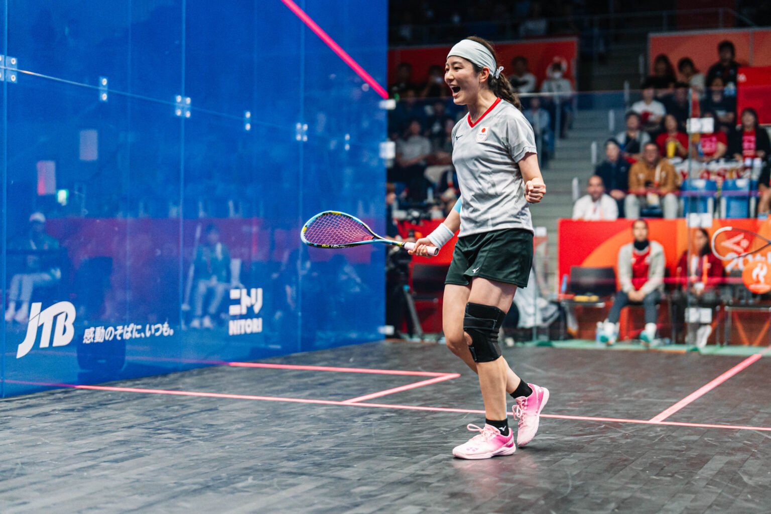 A squash player celebrating a point next to the blue glass wall of the show court at the Aichi-Nagoya 2026 Asian Games