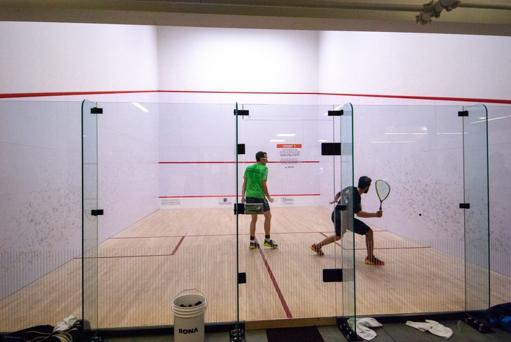 Two squash players moving around the T during a rally, one striking the ball while the other gives them room to swing