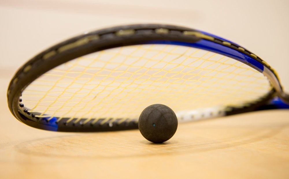 Strung squash racket and a double-yellow-dot squash ball resting on the wooden floor of a squash court