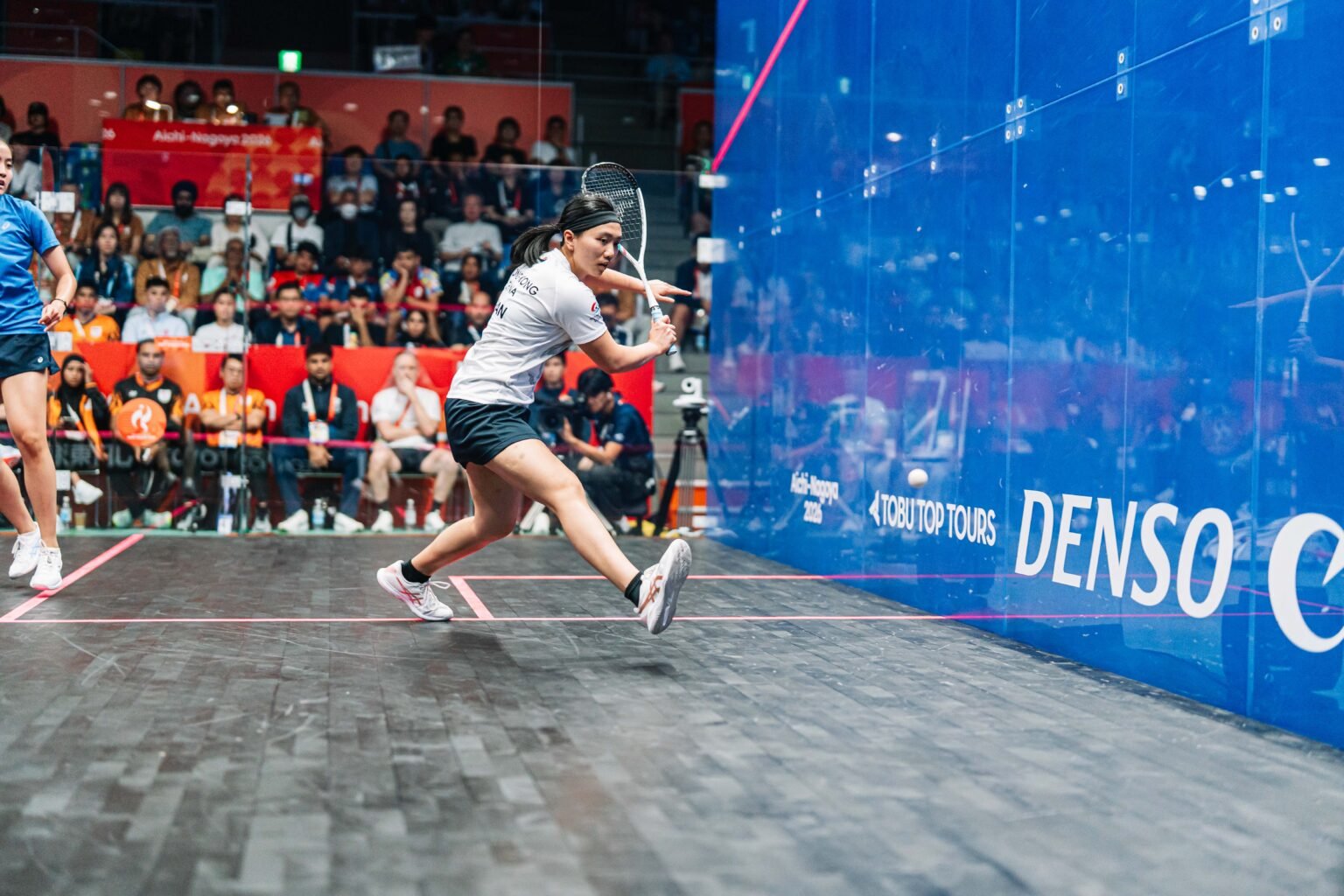 A squash player driving the ball tight along the glass side wall, the ball visible just off the wall as she swings through on the forehand