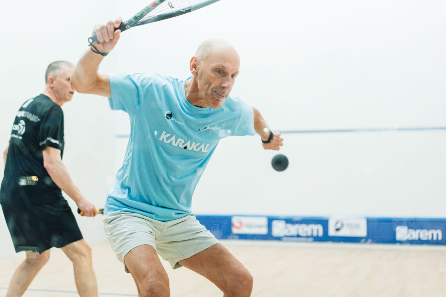 A club squash player mid-rally with the racket cocked high and ready and the ball in the air in front of him, prepared to strike before the bounce