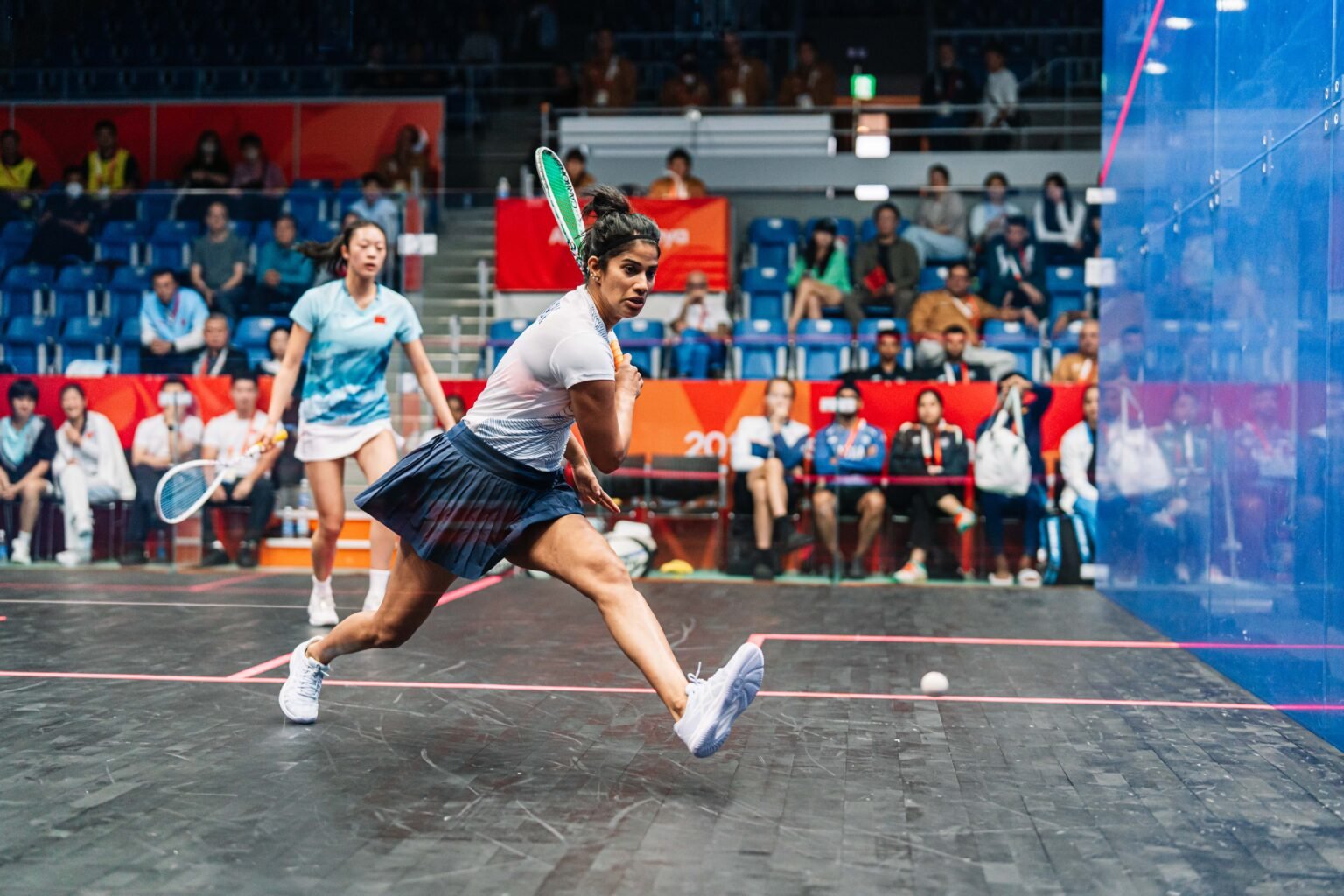 A women's singles player lunging forward to reach a low ball during a rally on the glass court at the Aichi-Nagoya 2026 Asian Games