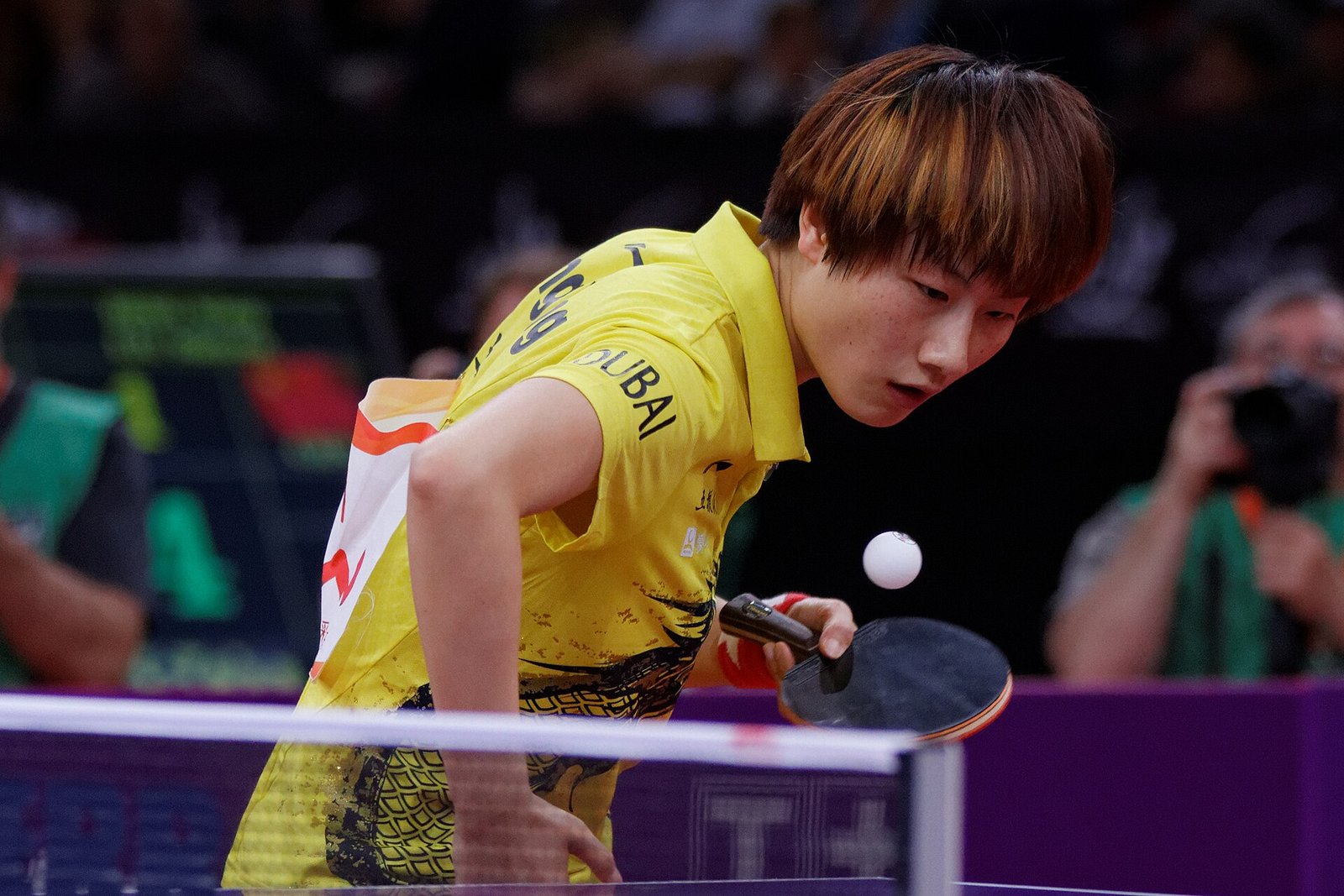A player leaning low over the table about to brush the ball with an open backhand face just above the surface, the ball hovering at racket height