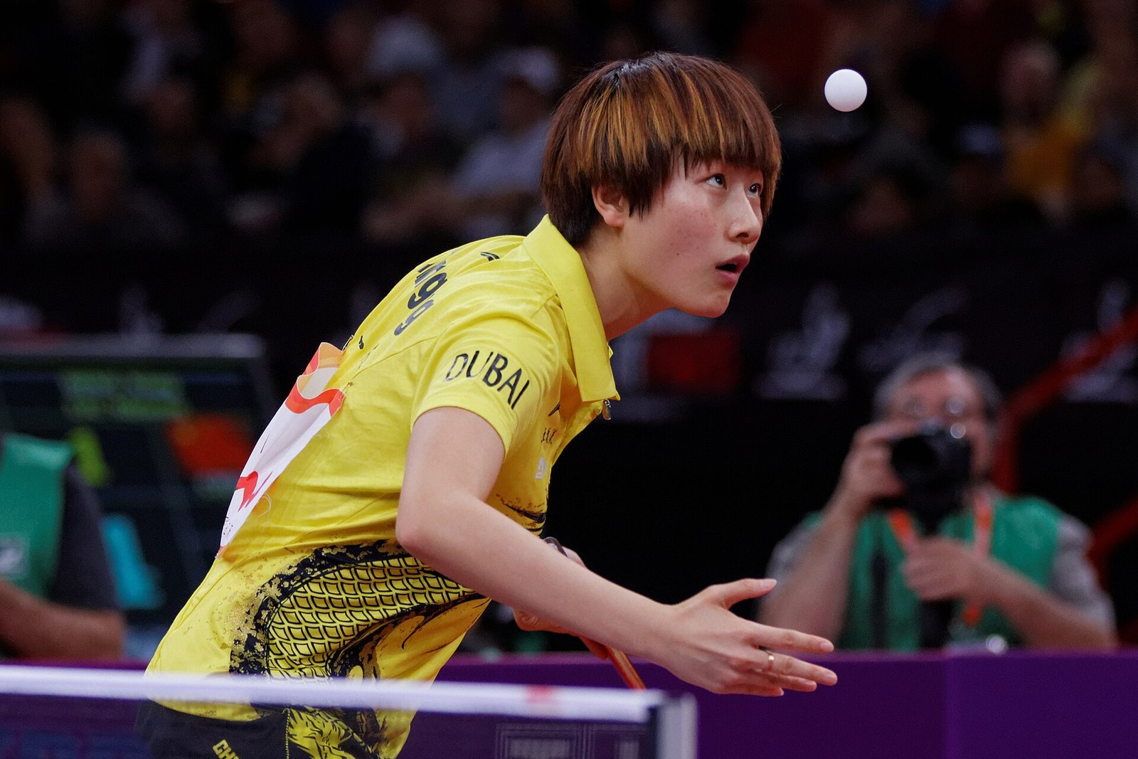 A player tossing the ball up with an open free hand to serve backhand, racket drawn back low on the backhand side, eyes fixed on the ball above the net