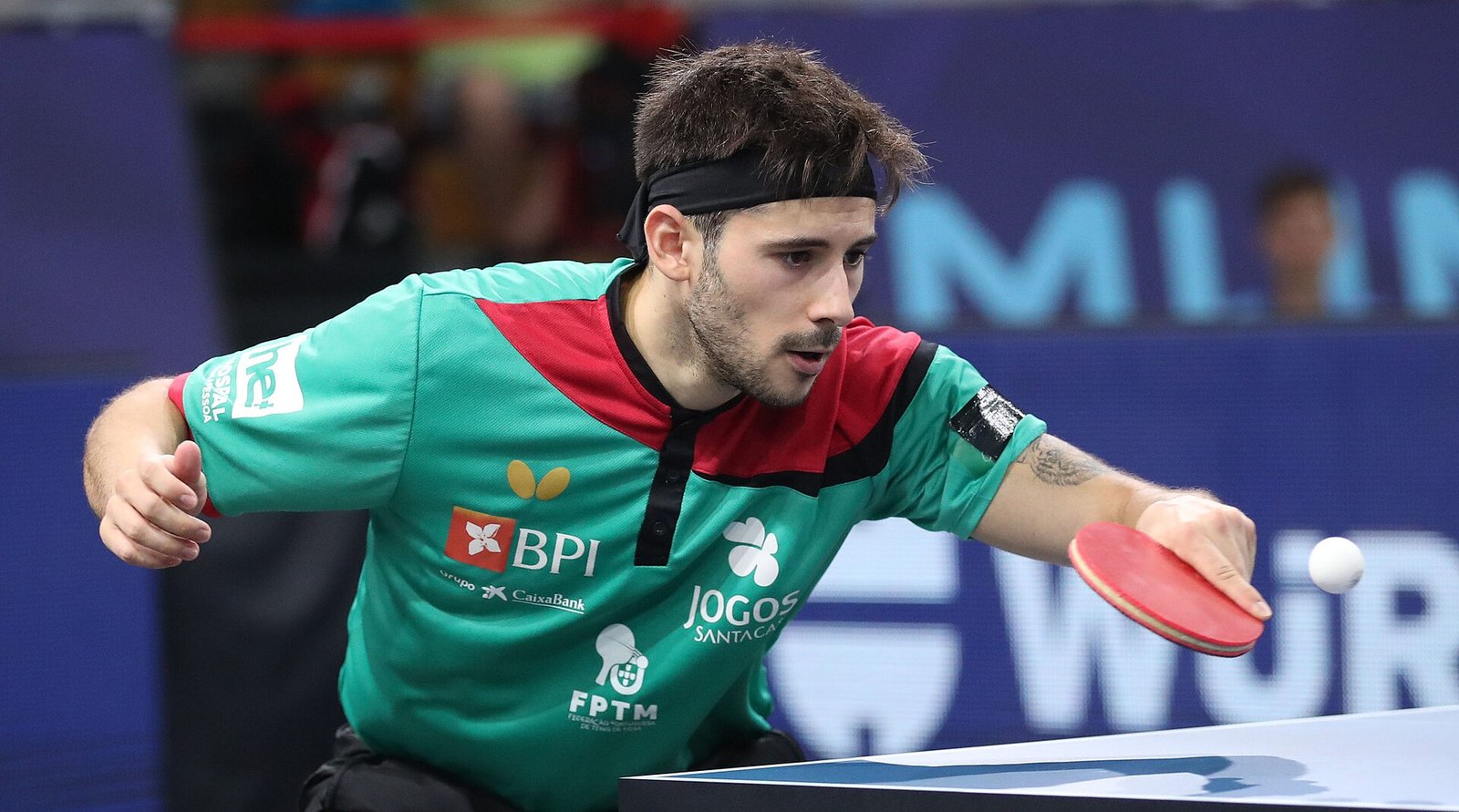 A player playing a backhand stroke close to the table, the red bat meeting the white ball just in front of his body with his free arm back for balance