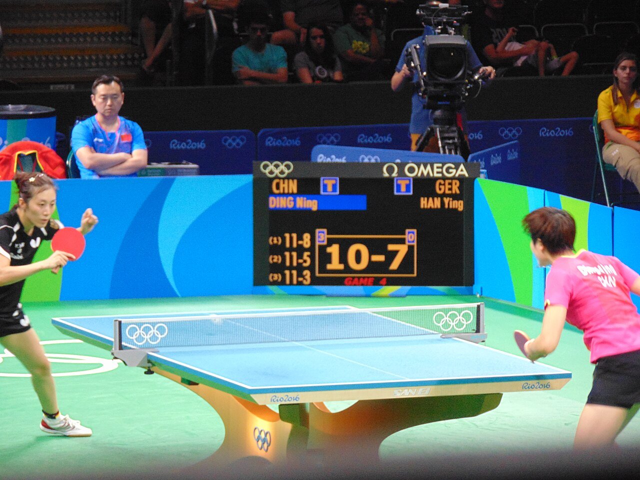 Two table tennis players reset in ready positions at opposite ends of the table between shots during a match at the Rio 2016 Olympics, the scoreboard behind them