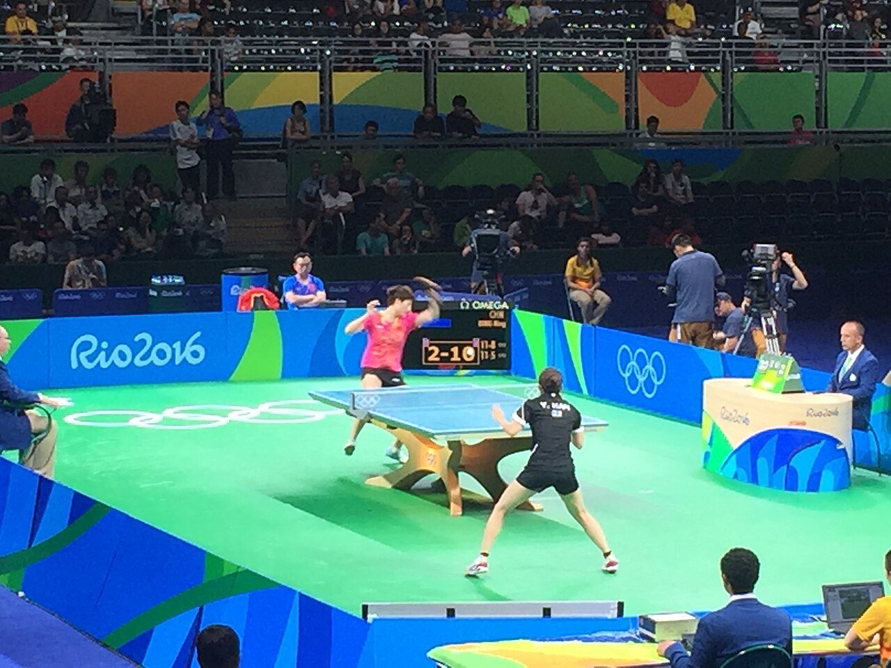 Wide view of an Olympic table tennis rally with one player in a wide ready stance in the foreground and her opponent mid-stroke at the far end, both players' feet clearly visible on the court
