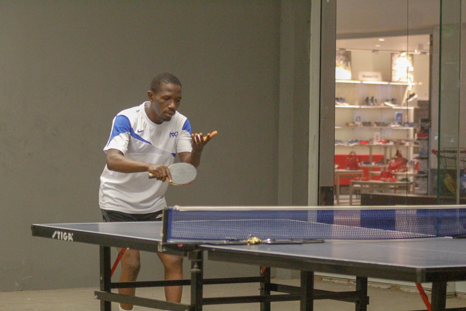 A table tennis player standing behind the table in a club hall, bat held in his playing hand and the ball in his free hand, ready to start a rally