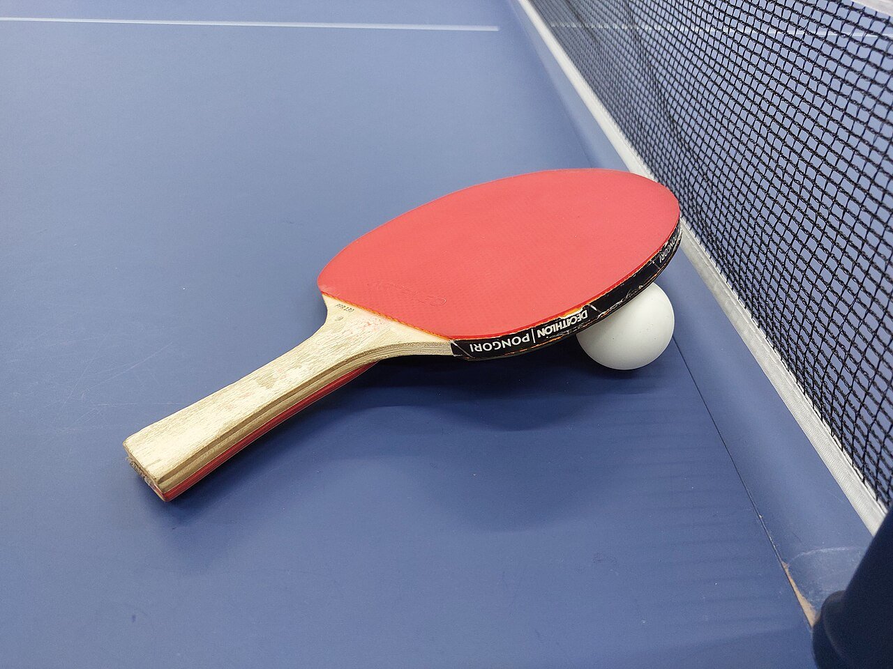 A table tennis racket and ball resting on the table beside the net — black rubber on one face, a bright colour on the other