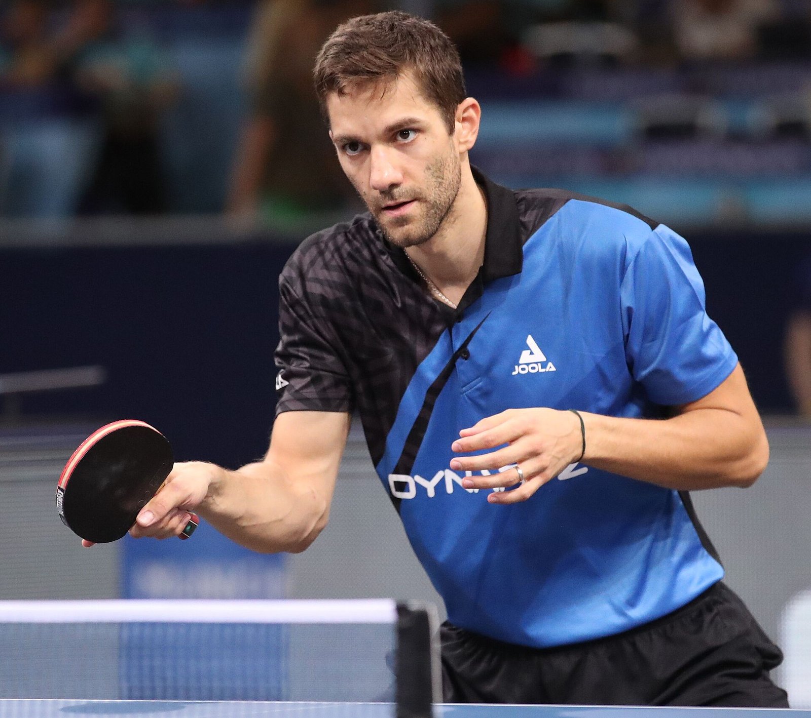 A table tennis player in the ready position behind the table, knees bent and weight forward, bat held out in front of his body as he watches the ball