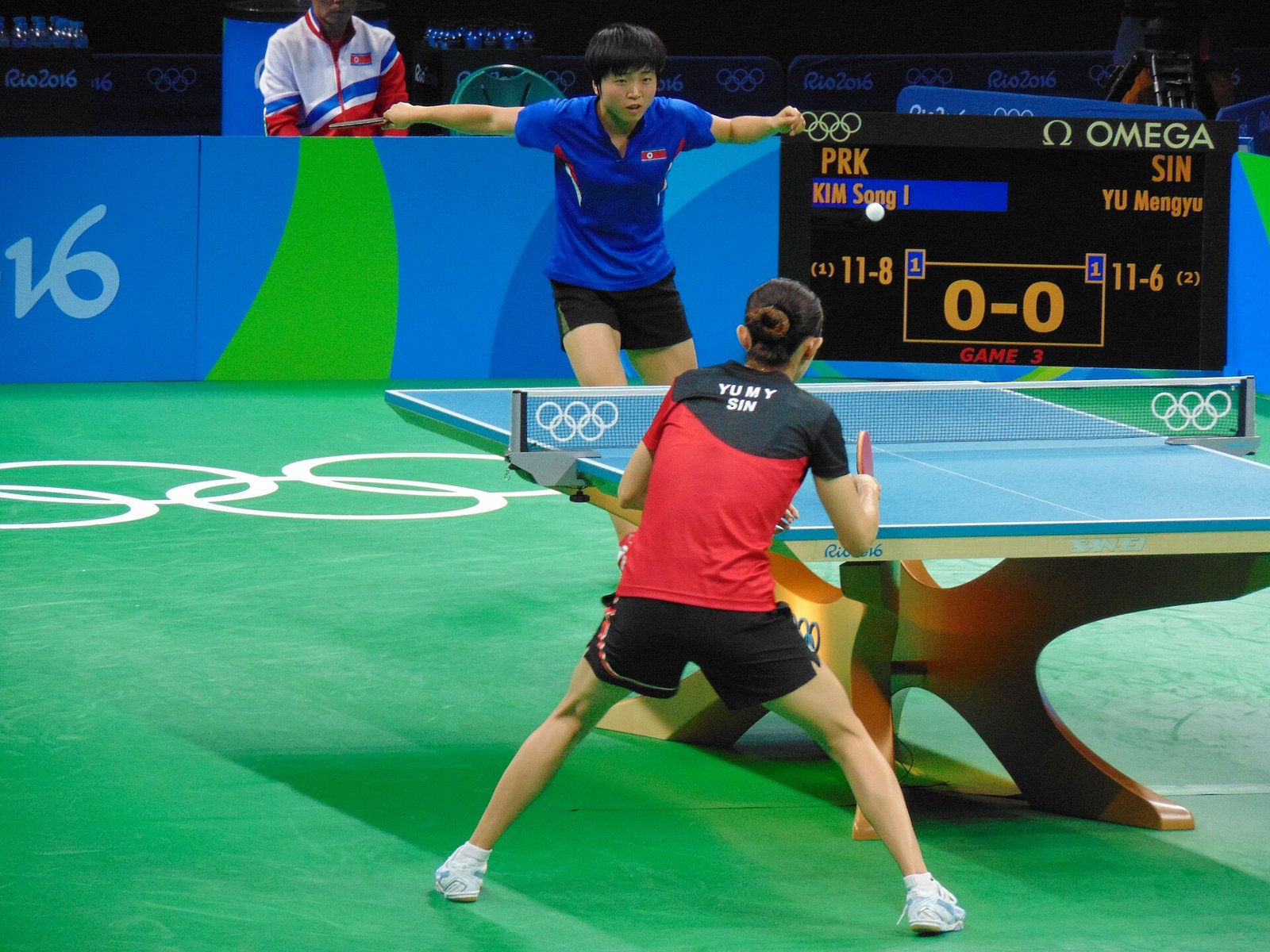 A wide view of an Olympic table tennis match at the start of a point, the server delivering from one end while the receiver waits in a low ready position at the other