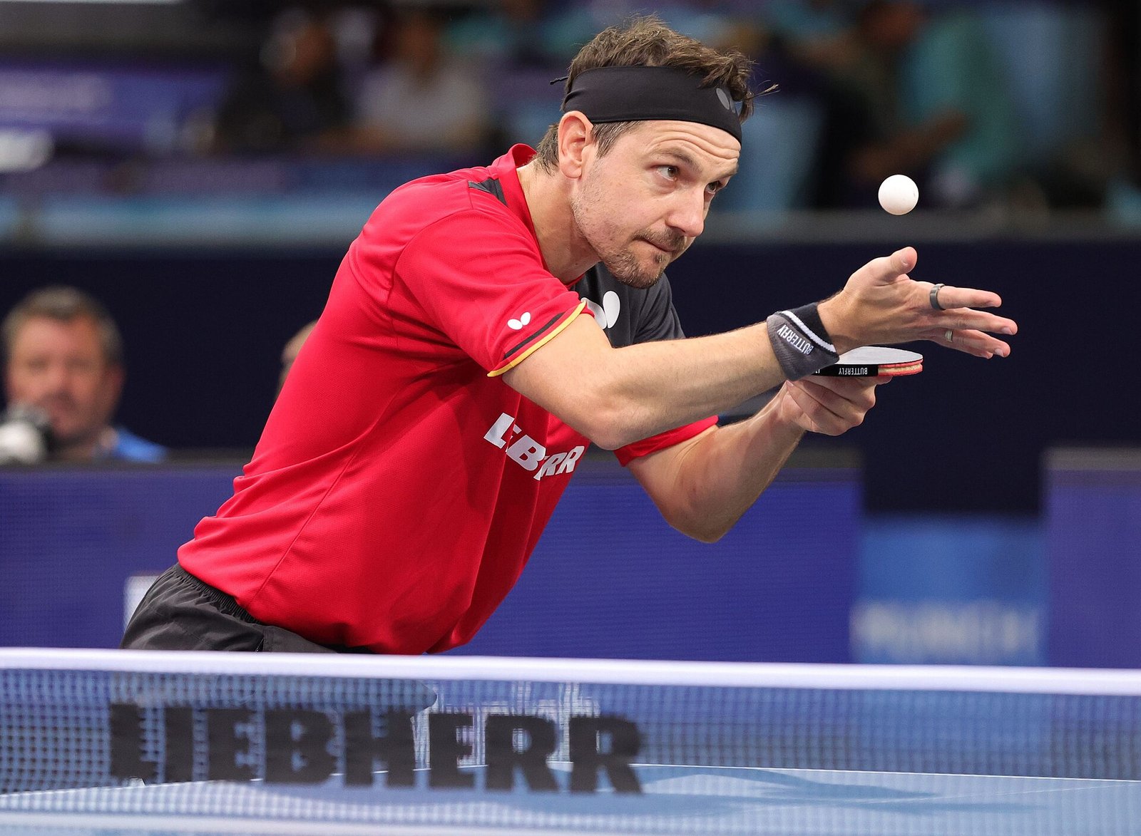 A player at the start of the serve, the ball tossed into the air from his open free hand while his bat waits in the other hand, the net in the foreground