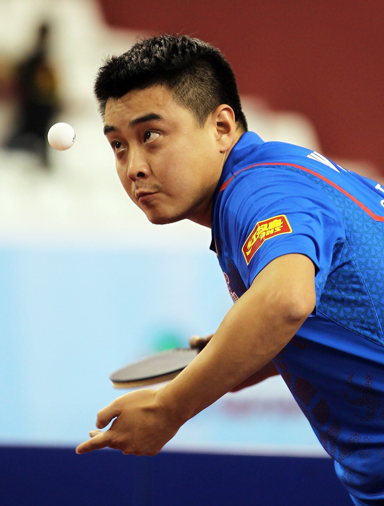 A penhold player a moment after tossing the ball for a serve, eyes on the ball as it hangs in the air, racket held low and ready behind him