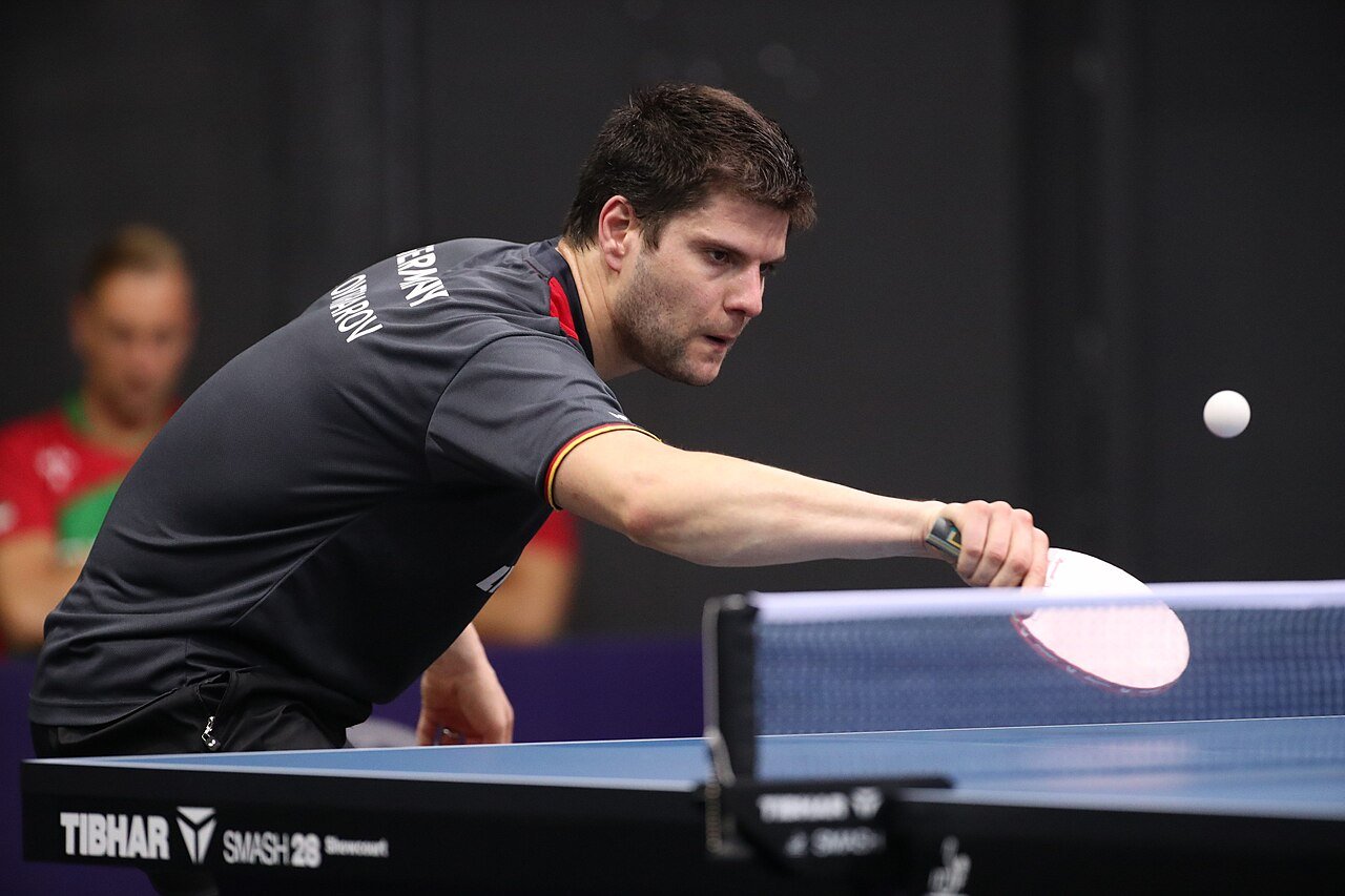 Table tennis player stepping in close to the table and leaning forward over it with his racket extended to reach a short ball at the European Championships